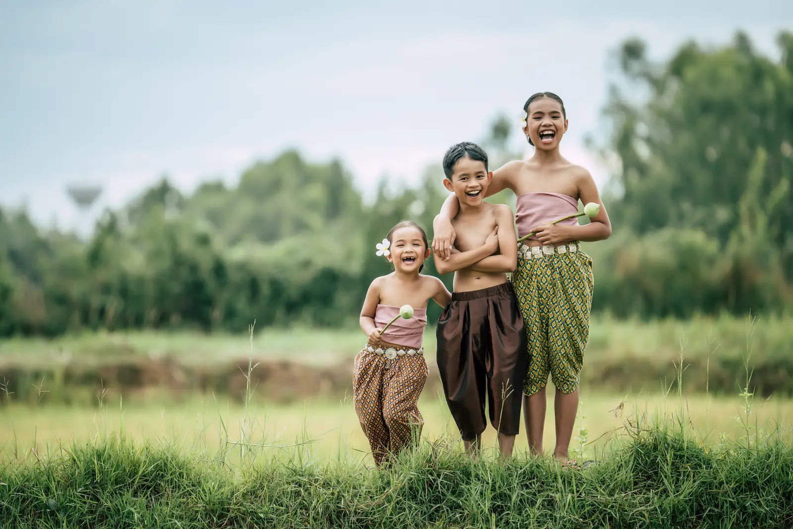 Three laughing children in traditional Thai dress stand at the edge of a green field, the two girls holding lotus buds.
