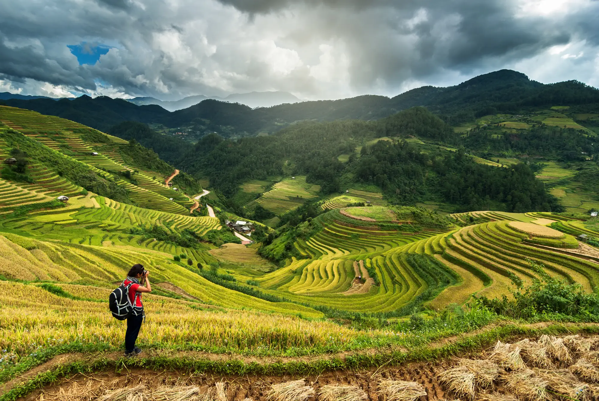 A walker with a backpack photographs terraced rice fields curving down a green valley under heavy cloud.