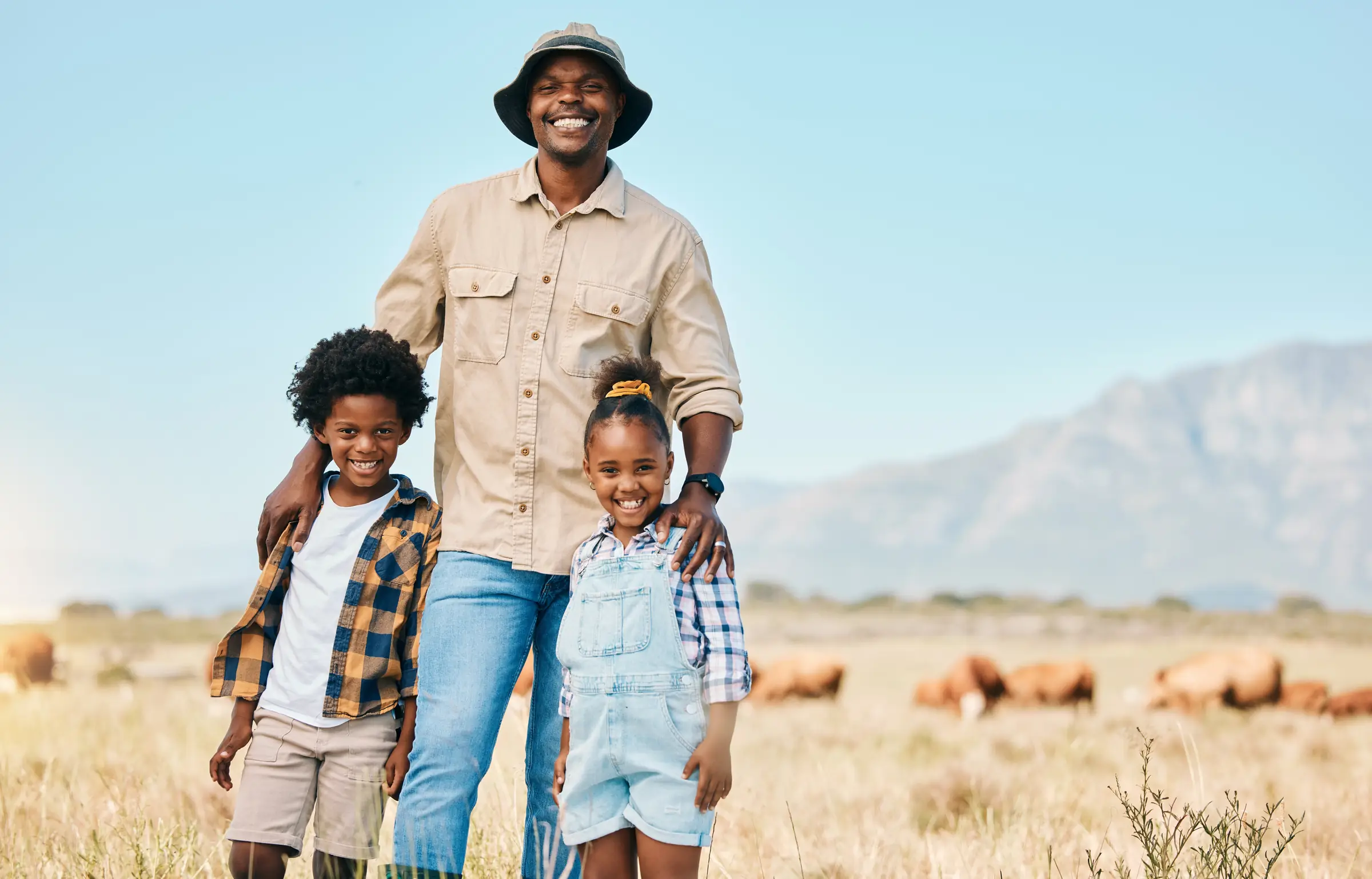A smiling father in a bucket hat stands in a sunlit field with his arms around his son and daughter, cattle grazing and mountains behind.