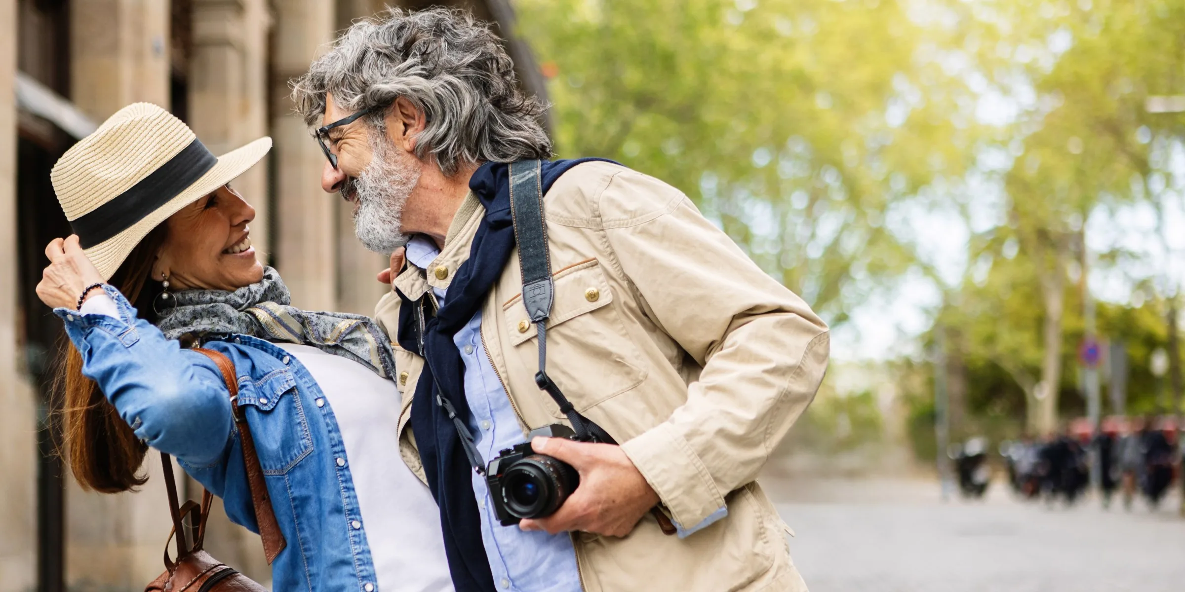 A couple in their sixties laughing together on a tree-lined city street.