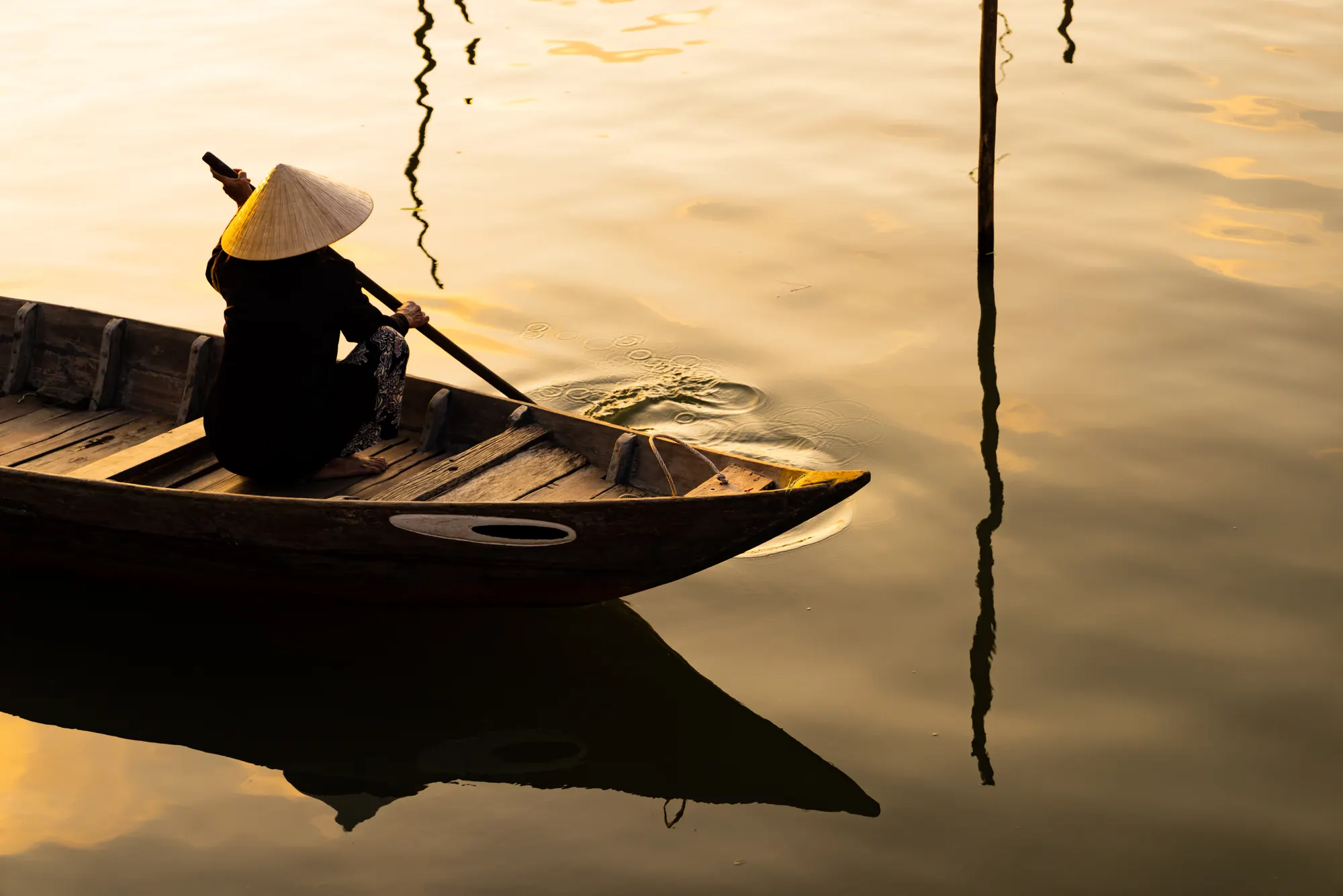 A rower in a conical hat paddles a wooden boat across still water at golden hour.
