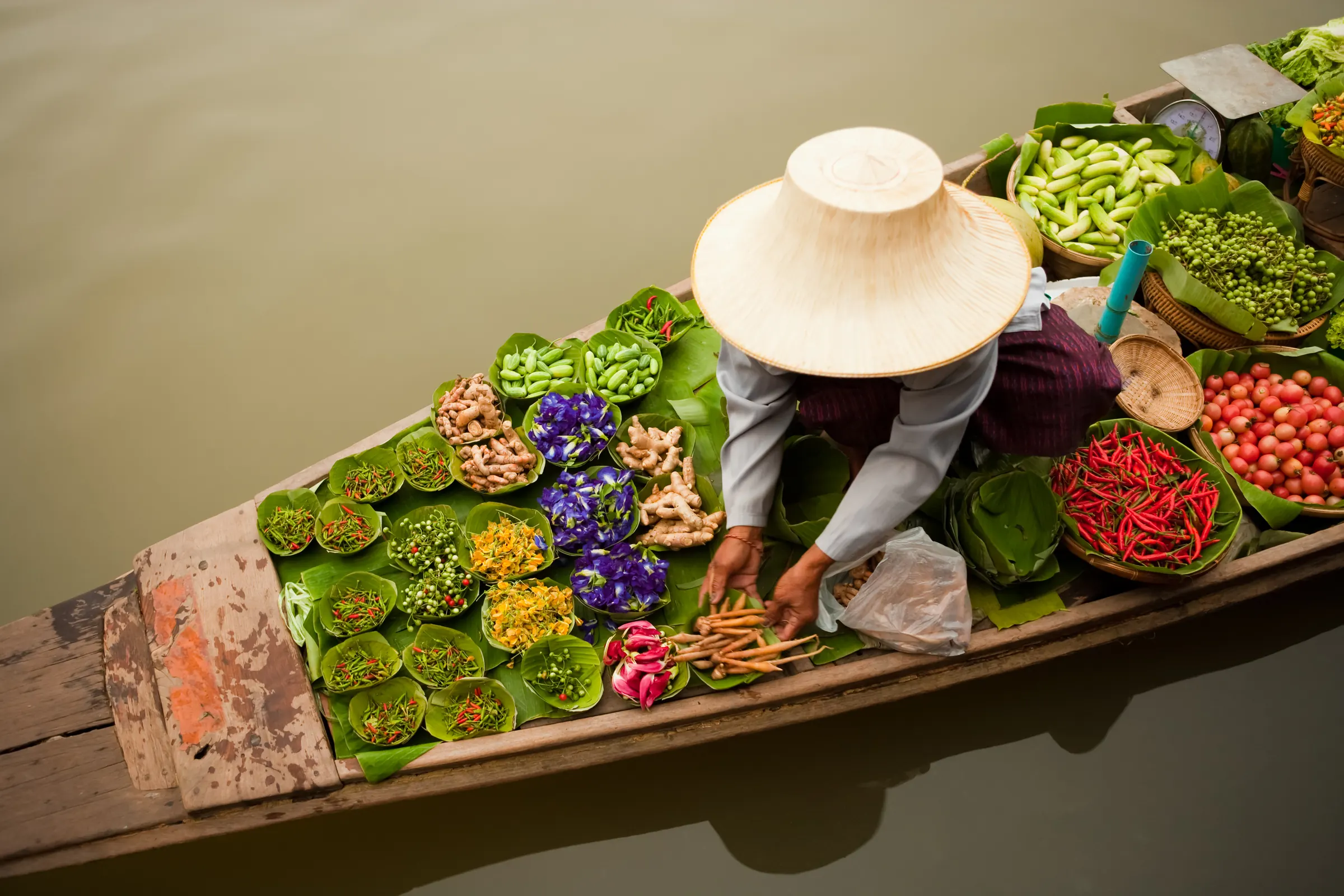 A vendor in a conical hat arranges vegetables and spices in a wooden boat at a Thai floating market.