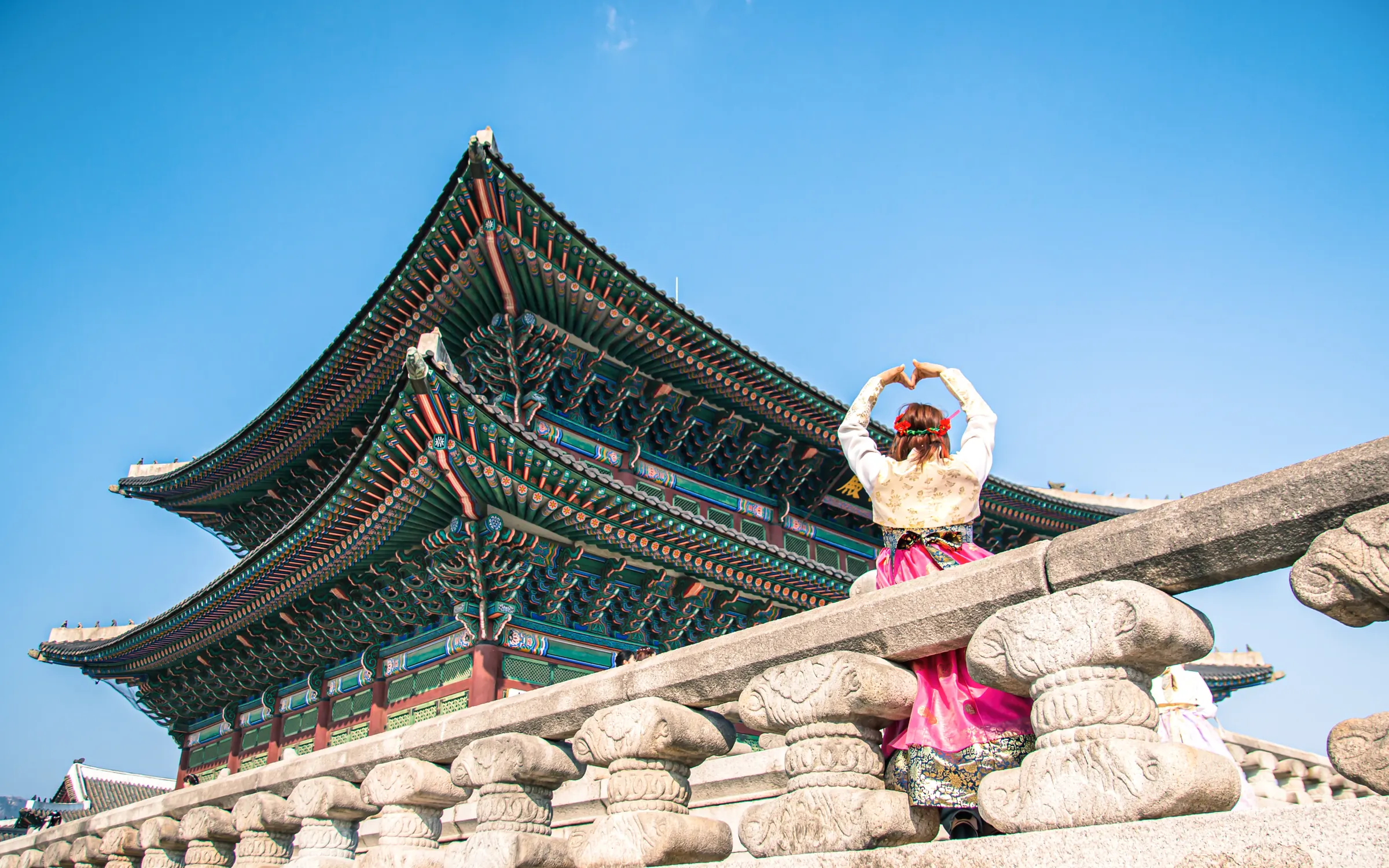 A traveler in a pink hanbok raises her arms in a heart shape on the stone terrace of Gyeongbokgung Palace in Seoul.