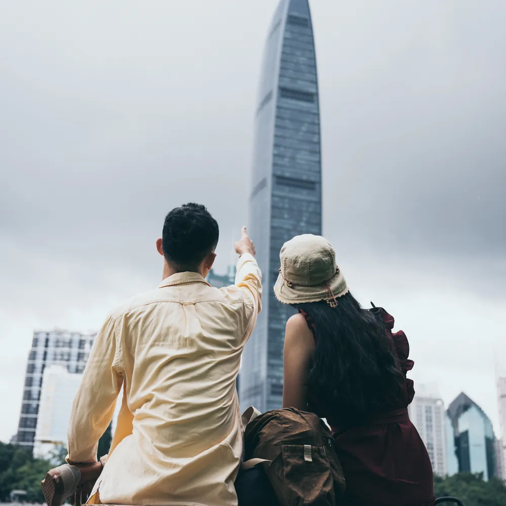 A couple seen from behind look up at a skyline of towers under a grey sky, one of them pointing.