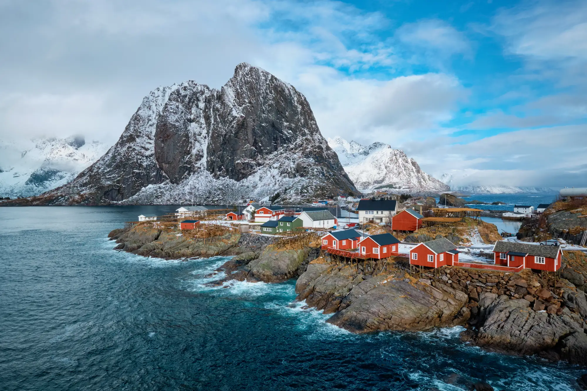 Red fishermen’s cabins on a rocky shore beneath snow-dusted peaks in the Lofoten Islands, Norway.
